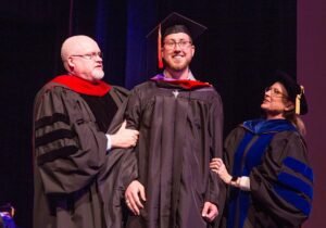 첥Ӱgraduate student at graduation standing between two professors smiling.